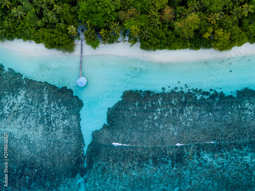 Aerial view of Kihaadhoo island with a wooden pier extending into the turquoise lagoon, surrounded by coral reefs and lush palm trees in Kihaadhoo, Baa Atoll, Maldives.