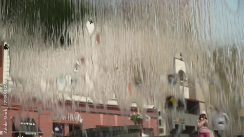 A dense, continuous stream of water flows from the edge of a fountain, shot from inside, with an old architectural building in the background, in the city center, on a hot summer day.