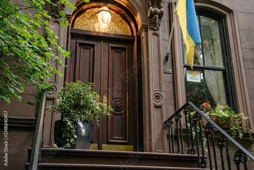 A Ukrainian blue and yellow flag hangs beside an ornate wooden doorway of a historic New York brownstone townhouse. Decorative iron railings, stone facade, warm lantern light, and green trees frame