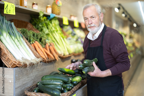 Photography Elderly man chooses ripe zucchini for purchase in greengrocer shop