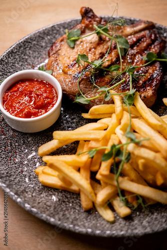 Close-up of a juicy grilled bone-in steak with dark sear marks, served with golden French fries, spicy red dipping sauce and fresh microgreens on a textured ceramic plate.