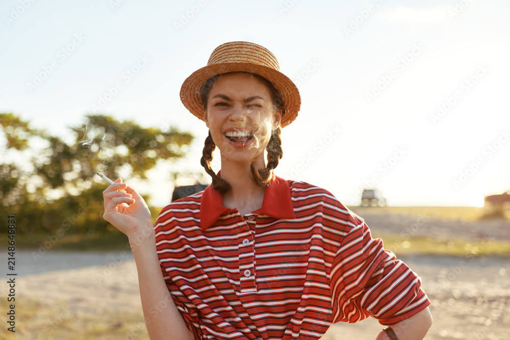 Obraz premium A joyful young woman with braided hair and a straw hat smokes a cigarette outdoors. The authentic lifestyle scene captures a relaxed moment of freedom in a sunny summer, creating a vibrant mood.