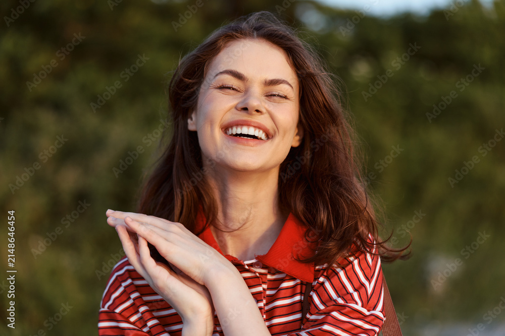 Obraz premium A cheerful woman outdoors in a red striped shirt smiles with eyes closed, enjoying a sunny moment. A candid lifestyle scene highlighting happiness, relaxation, and natural joy.