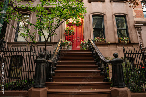 Wide view of a historic New York brownstone townhouse featuring stoop stairs, red door, black iron fence, and stone facade. A leafy tree and flower boxes enhance the urban residential streetscape.