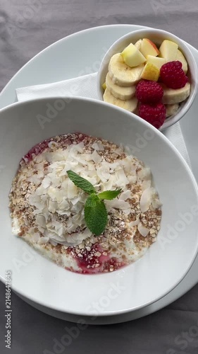 close up top down view of plated breakfast bowl with yogurt, granola, coconut flakes, mint, berry sauce, and mixed fruit on clean ceramic table in soft daylight