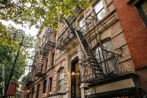 Historic New York brownstone apartment building with classic brick facade and black metal fire escapes viewed from street level under green trees. Urban city architecture with detailed windows