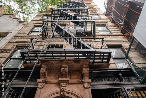 Dramatic low angle perspective of black metal fire escape attached to a historic New York brick building facade. Urban architectural detail highlighting exterior staircase, windows, and classic