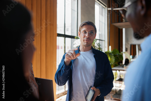 Young man with glasses holding a digital tablet, discussing ideas and collaborating with diverse colleagues during a creative business meeting in a modern office space