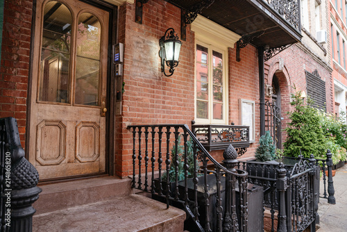 Traditional New York brownstone entrance with wooden door, iron railing, and brick facade along a quiet residential street. Historic Manhattan architecture featuring detailed exterior design and urban