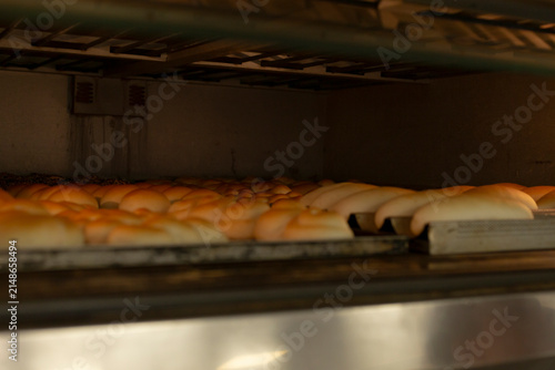 rows of golden bread rolls rise and brown on metal conveyor belt inside dark industrial oven, with low warm light, steel racks, and fresh baked texture during batch