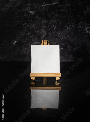 Vertical photo mock up of small square blank white canvas sitting on a mini wooden art easel sitting on black mirror reflective surface and dark background.