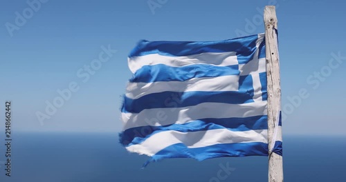 Greek flag on a sun‑bleached wooden stick whips in fierce wind high above the Aegean Sea on Crete, with bright blue water and rugged coastal terrain visible as the camera captures the dramatic motion.