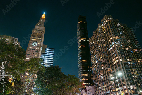New York City skyline featuring historic clock tower and modern residential and office skyscrapers illuminated at night. Tall buildings rise above trees and streets creating a dramatic Manhattan