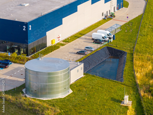 Aerial view of an industrial warehouse with a large metal water tank, parking lot, and retention pond in Kowale, Pomeranian Voivodeship, Poland.
