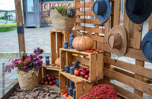 Rustic autumn themed photo zone with a pumpkin, apples, wicker baskets of flowers, and cowboy hats on wooden pallets. A vibrant harvest display at an outdoor street festival