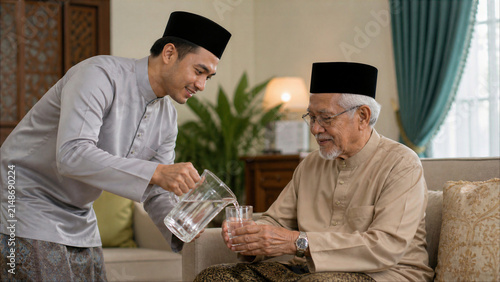 Young Malay man in traditional Baju Melayu pouring water for his elderly father at home, showing respect and filial piety.