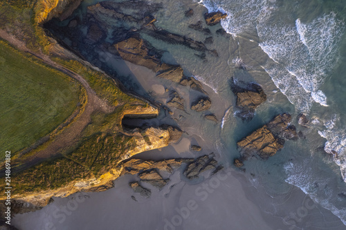 Aerial view of As Catedrais beach featuring dramatic rock formations, sandy shoreline, and turquoise waves crashing against the cliffs in Spain.