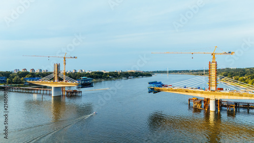 Aerial view of a bridge under construction over the Danube River with tall cranes and concrete pillars rising above the water in Novi Sad, Serbia.