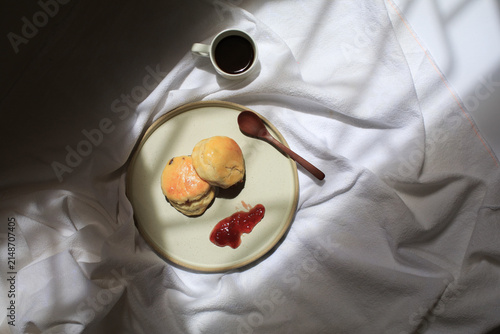 Scones with dollop of jam beside steaming cup of coffee, bathed in dappled sunlight. Freshly baked in morning.
