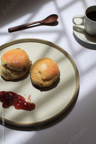 Scones with dollop of jam beside steaming cup of coffee, bathed in dappled sunlight. Freshly baked in morning.