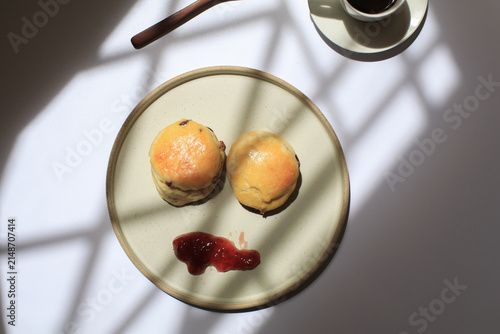 Scones with dollop of jam beside steaming cup of coffee, bathed in dappled sunlight. Freshly baked in morning.