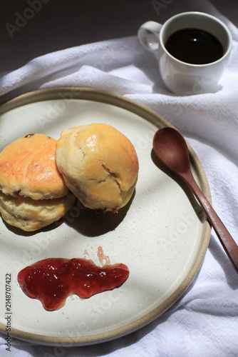 Scones with dollop of jam beside steaming cup of coffee, bathed in dappled sunlight. Freshly baked in morning.