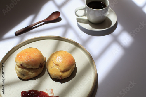 Scones with dollop of jam beside steaming cup of coffee, bathed in dappled sunlight. Freshly baked in morning.