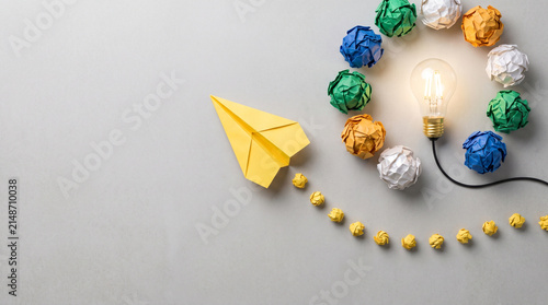 Creative innovation concept showing a yellow paper airplane flying towards a glowing lightbulb leaving discarded crumpled ideas behind