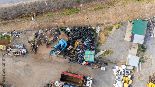 Aerial view of a scrap yard with a large pile of discarded tires, a blue excavator, a red dump truck, and industrial debris on a gravel lot in Sremska Mitrovica, Vojvodina, Serbia.