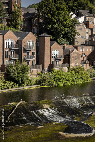 View of the River Wear weir with historic brick buildings and lush greenery along the riverbank in Durham, England, United Kingdom.
