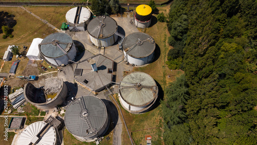 Aerial view of industrial storage tanks and facilities with some structures under maintenance or demolition, surrounded by lush green trees in Milan, Lombardy, Italy.