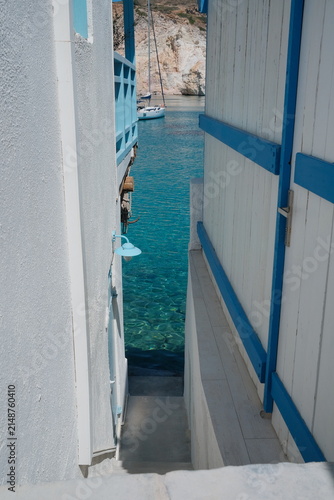 фотографія A narrow passage between whitewashed Greek buildings reveals vivid turquoise water beyond, combining blue accents, traditional island architecture and the bright colors of the Aegean