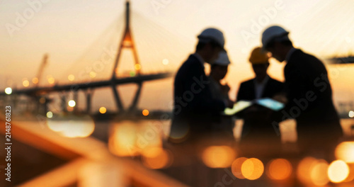 Silhouettes of construction professionals in hard hats discussing a project during sunset, with a bridge and city lights in the background.