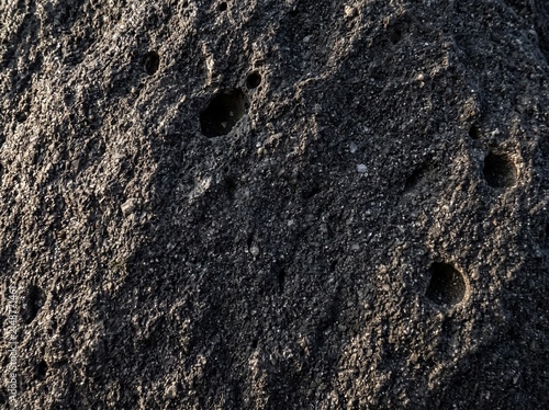 Dark basalt volcanic rock fills the frame with dense texture and vesicles in raking sidelight — volcanic geology as the darkest most dramatic fine art. Ideal for dramatic and dark design backgrounds.