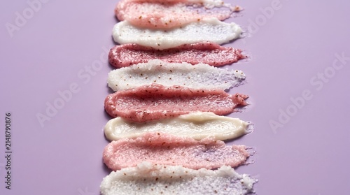 Swirls of colorful body scrub textures are artfully arranged in a vertical line against a soft pastel background, showcasing a variety of natural ingredients and gentle exfoliating properties.