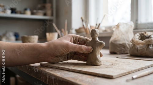A hand shaping a small clay figure on a wooden table. The workspace is cluttered with pottery tools and materials, illuminated by natural light from a window.