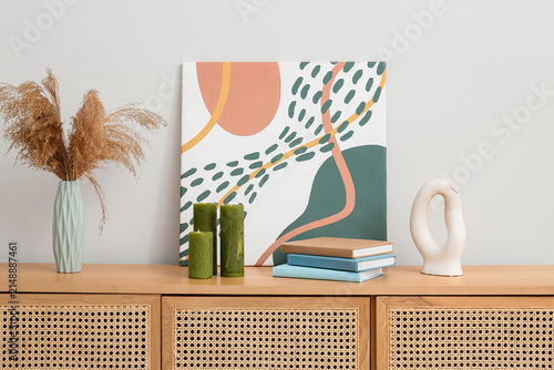 Vase with pampas grass, burning green candles, books and painting on commode near light wall