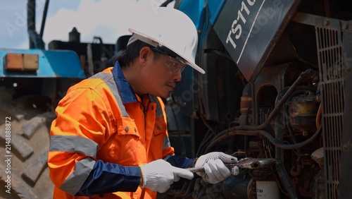 Asian male mechanic engineer in safety helmet repairing heavy tractor machinery engine with wrench tool outdoor. Professional industrial vehicle maintenance and agricultural equipment service.