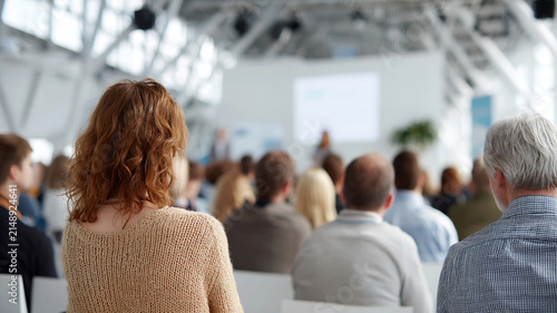 Corporate conference audience attendee seminar presentation, rear view of focused people seated in modern bright event hall
