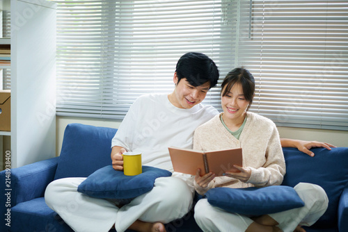 Фотография Young happy couple sits together on a cozy blue couch in their living room, looking at a memory book together and smiling while reminiscing about cherished times and drinking coffee