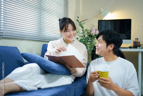 Konstfotografi Young happy couple sits together on a cozy blue couch in their living room, looking at a memory book together and smiling while reminiscing about cherished times and drinking coffee