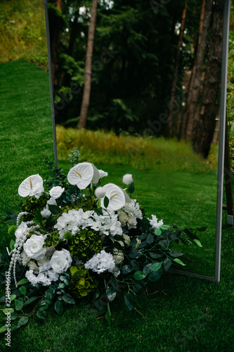A bouquet of various white flowers lies next to a large mirror on green grass against a background of a dense forest, decor for a wedding celebration