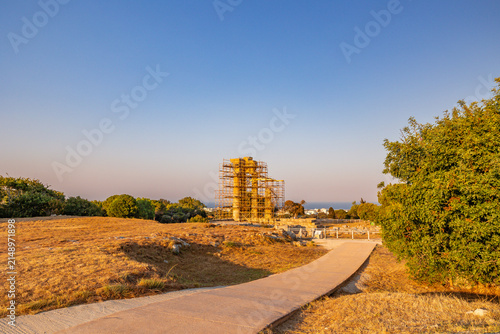 Ancient Temple of Pythian Apollo undergoing restoration on the Acropolis of Rhodes, Greece. Stone columns surrounded by scaffolding set against a clear blue sky during warm golden hour light.