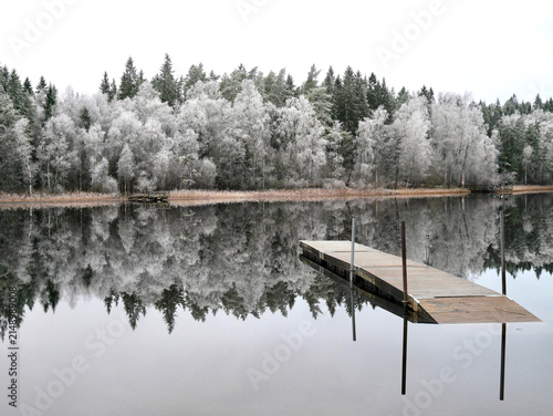 Wooden pier extending into a calm lake reflecting frost-covered trees on a quiet winter day in Sweden.