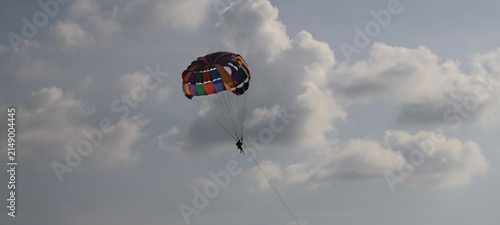 Принти на полотні A vibrant parasail canopy in rainbow colors carries a person high above the ocean, suspended by cords and tow line against a partly cloudy sky