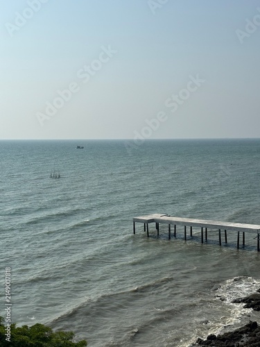 A peaceful seascape at Khao Sam Muk, Chonburi, Thailand, featuring gentle waves, a concrete pier, and a fishing boat on the horizon beneath a clear blue sky.