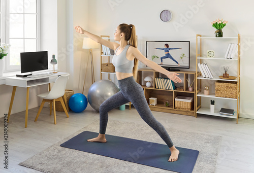 Woman practicing yoga workout at home television online. Full length female on a yoga mat mirrors a screen instructor for balance, flexibility and fitness. Concept shows mindful home exercise