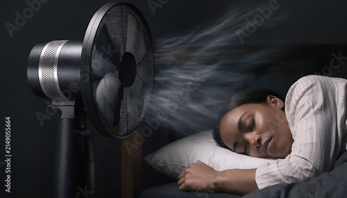 Young Black woman sleeping peacefully in a comfortable bed during a hot summer night, refreshed by the gentle breeze of a modern pedestal fan in a cozy dark bedroom.