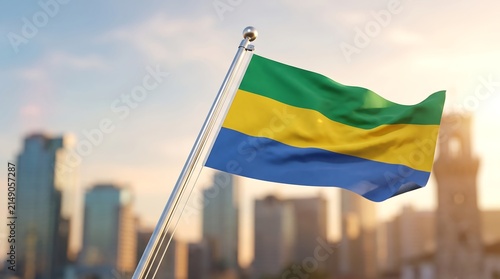 Gabon national flag waving on a silver flagpole against a blurred city skyline at sunset