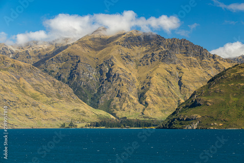 View of the mountains rising above the deep blue waters of Lake Te Anau under a clear sky with scattered clouds in Te Anau, Southland Region, New Zealand.